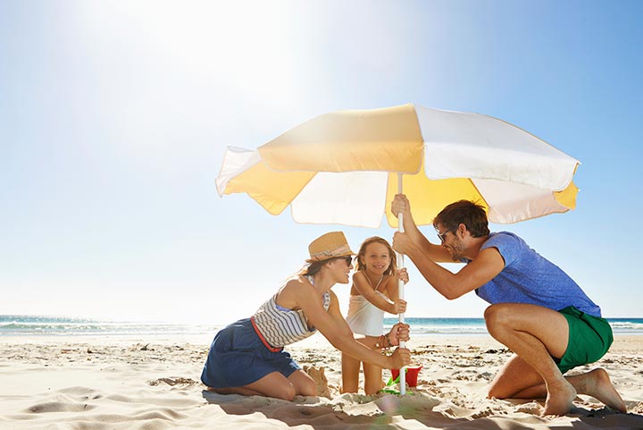 Foto: Eltern stellen mit Kind am Sandstrand einen Sonnenschirm auf.